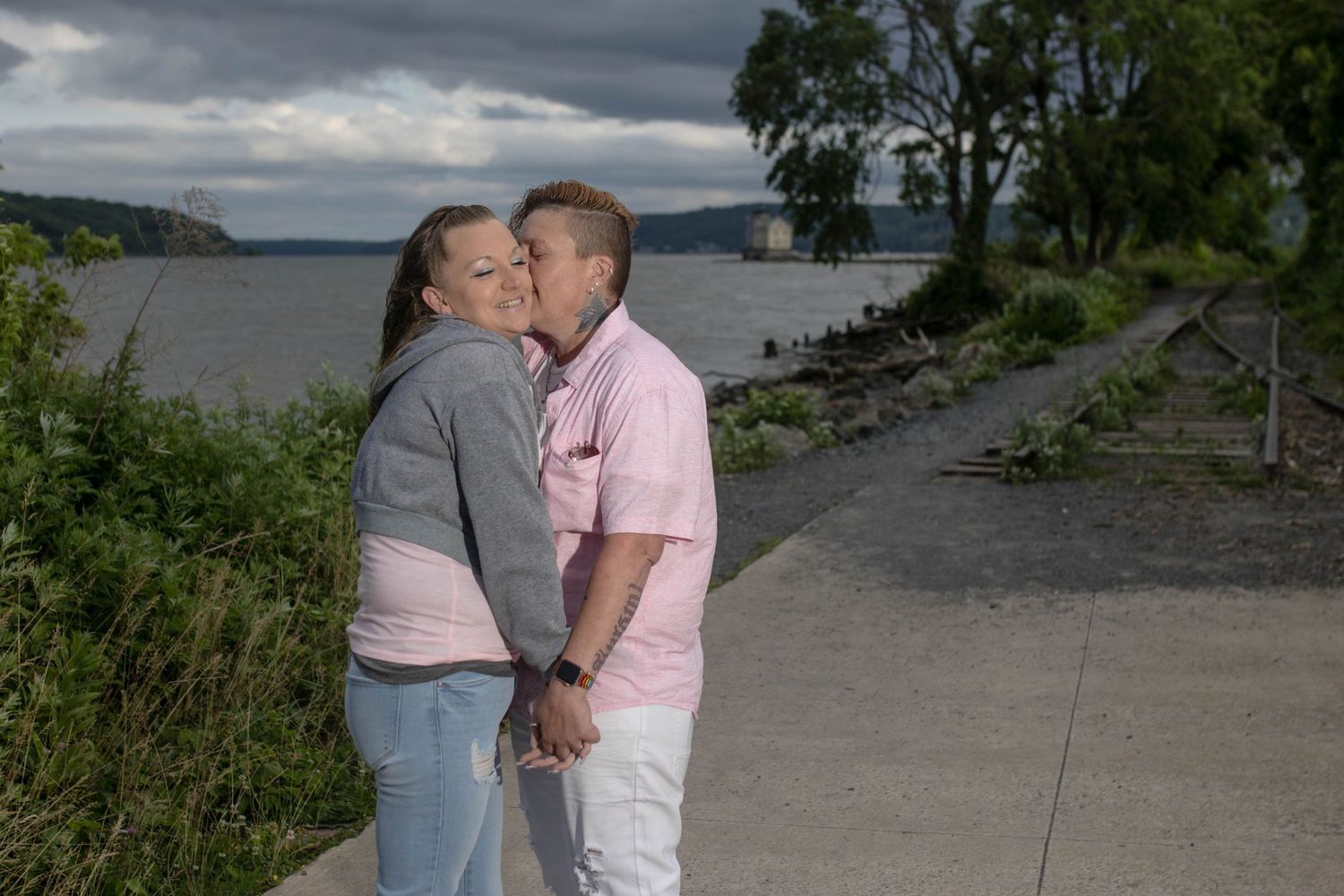 Couple shares intimate moment on waterfront path under cloudy sky.
