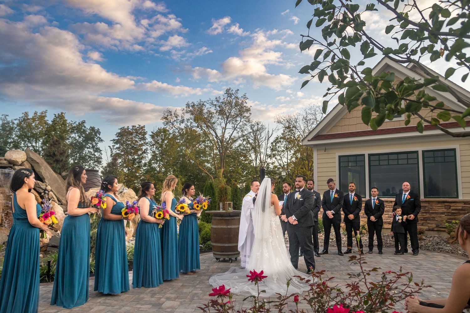 Wedding party in teal blue dresses and dark suits pose together outdoors under blue sky.