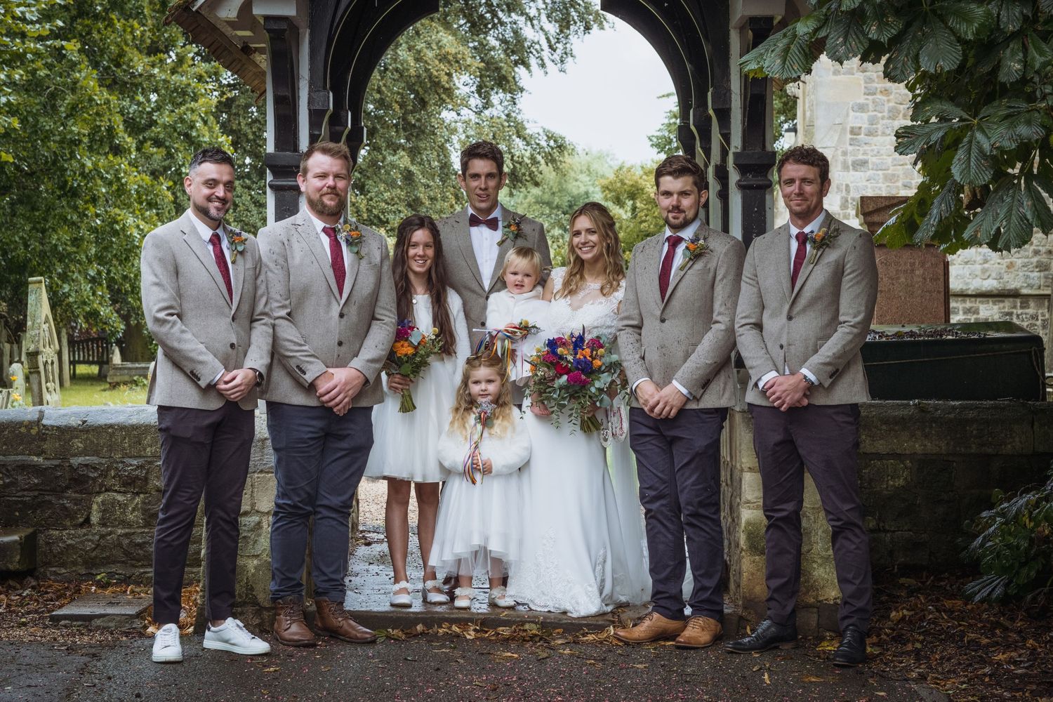 Wedding party in gray suits poses for a group photo under a stone archway.