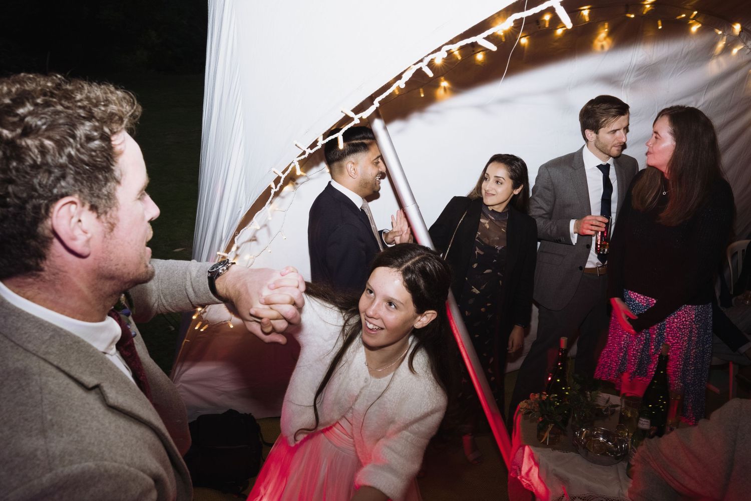 Wedding guests dancing and socializing under string lights in a tent.