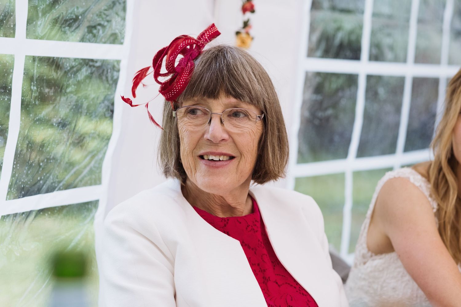 A guest in red attire and matching fascinator sits at a wedding reception.
