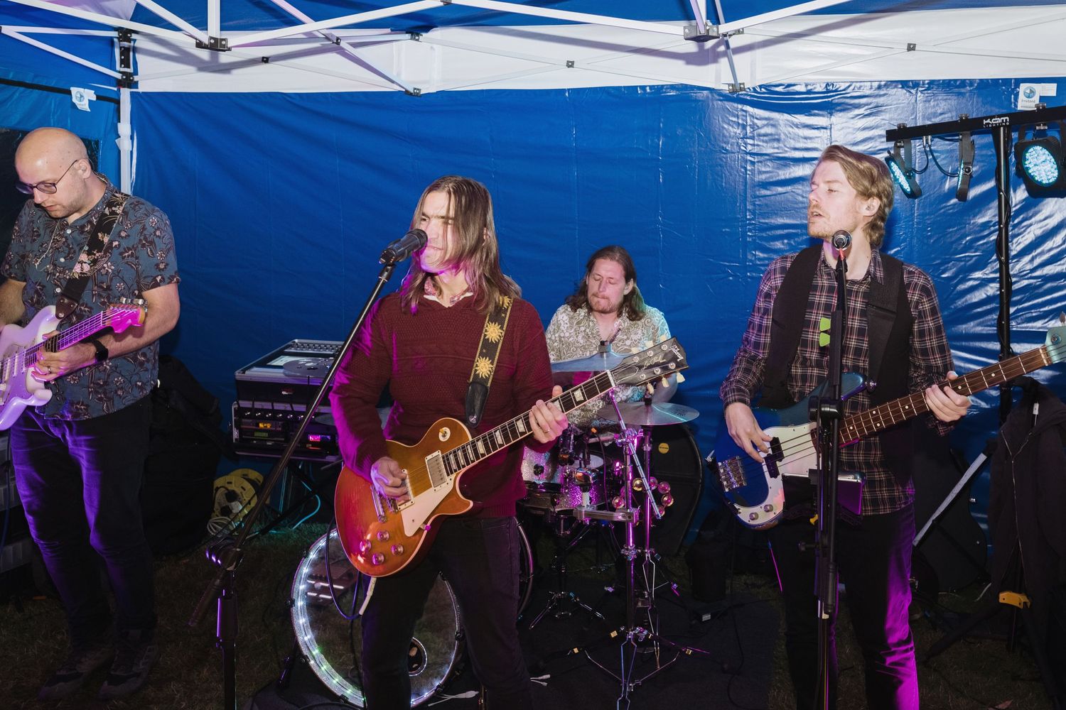 A band performs with guitars and drums under a blue tent with vibrant lighting.