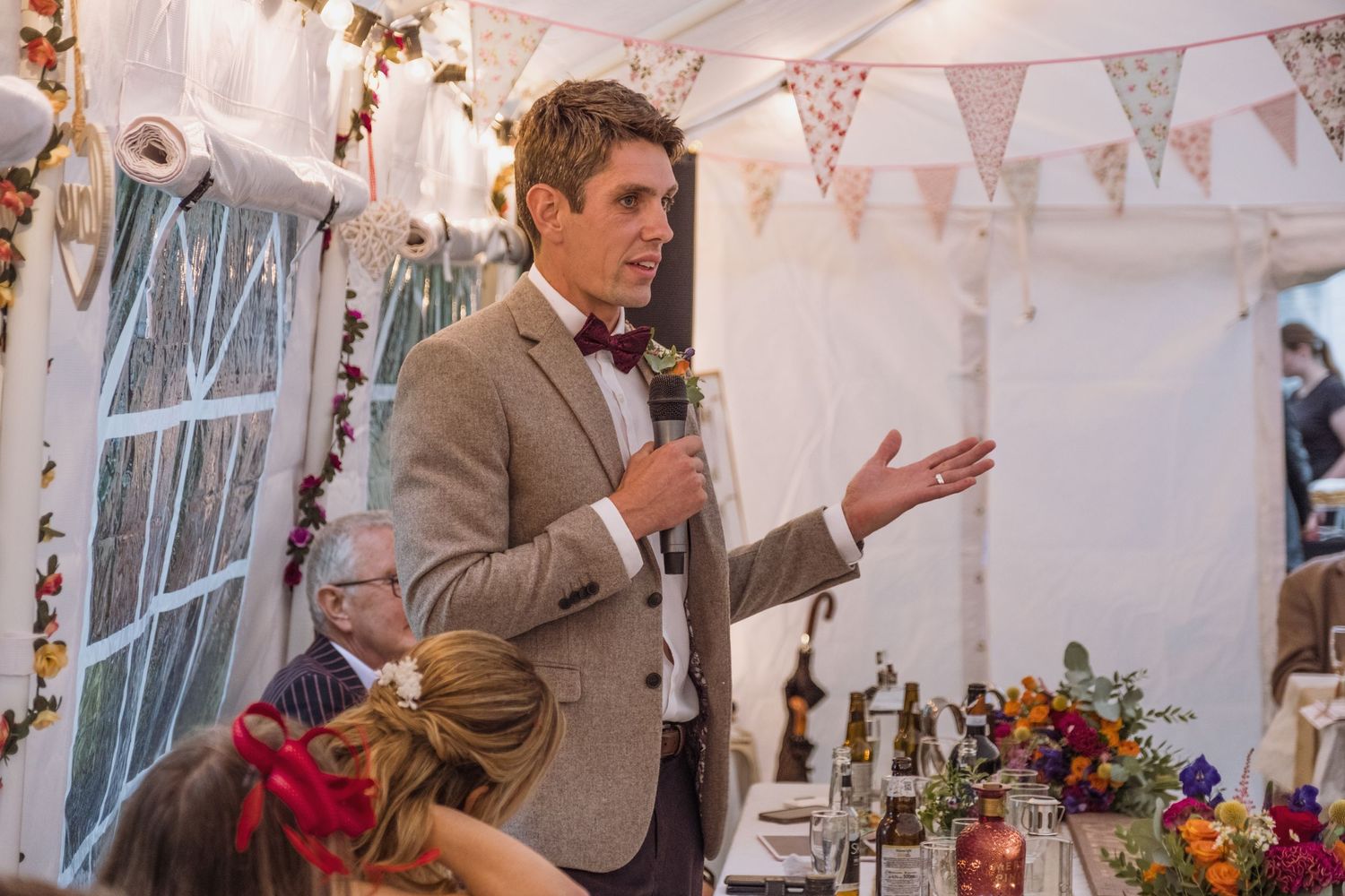 A speaker gives a wedding toast in a decorated tent with bunting and festive decor.