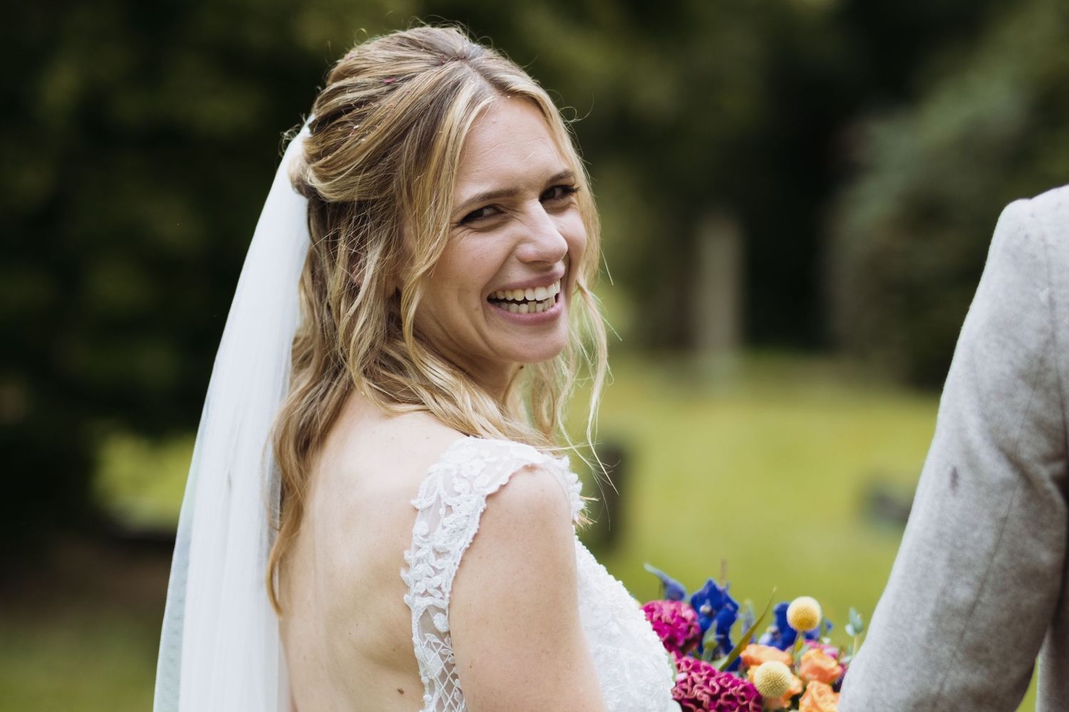 A bride in a lace dress smiles while holding a colorful bouquet outdoors.