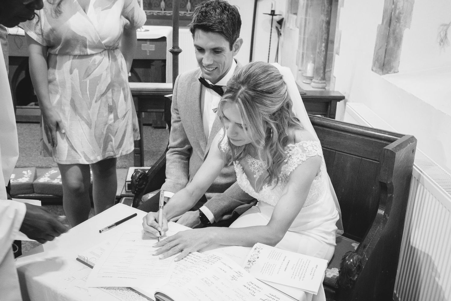 A couple signs documents together at a registration desk while a person in white stands nearby.
