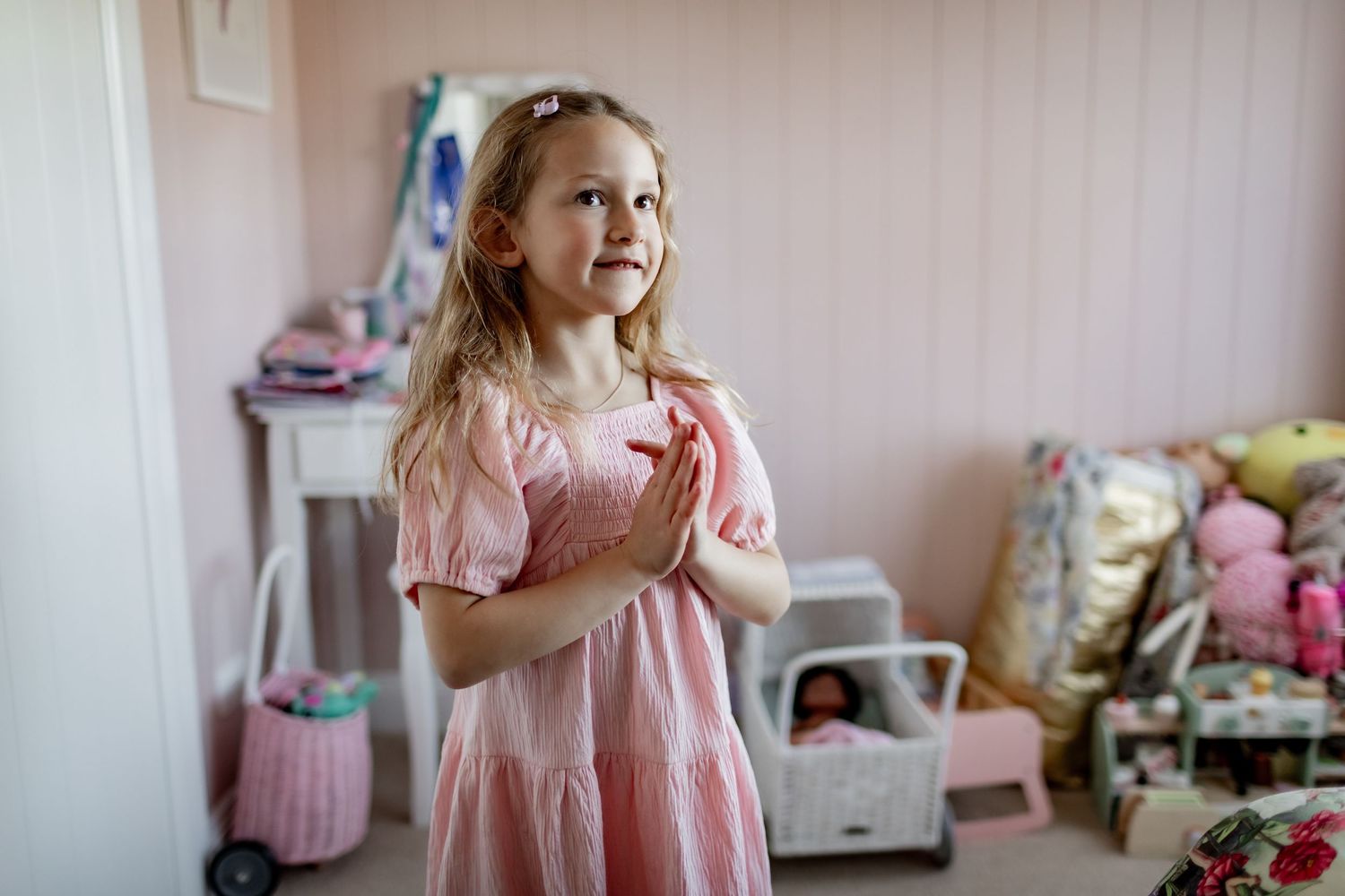 Child in pink dress standing in bedroom with toys and storage bins visible in background.