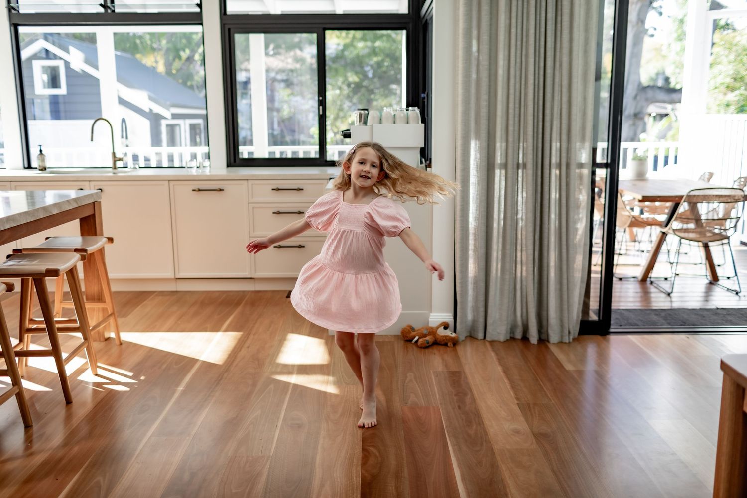 Young child in pink dress twirling and dancing joyfully on hardwood floor in sunlit room.