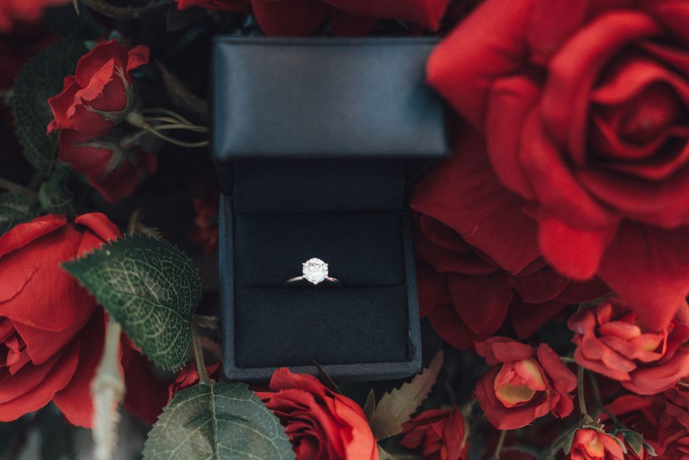 A diamond engagement ring in black box surrounded by vibrant red roses.