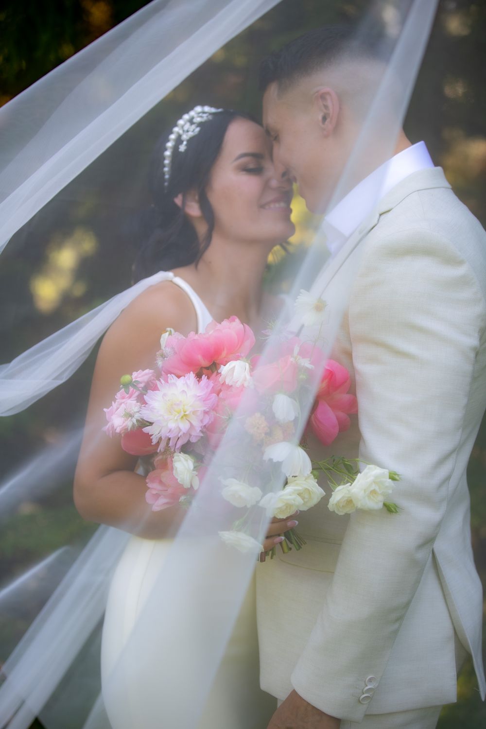 Intimate wedding moment captured through flowing veil with pink floral bouquet.