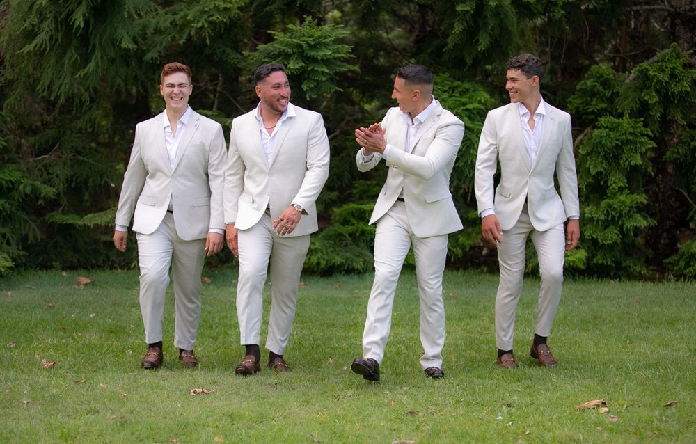 Groomsmen in matching white suits walking and laughing on green lawn.