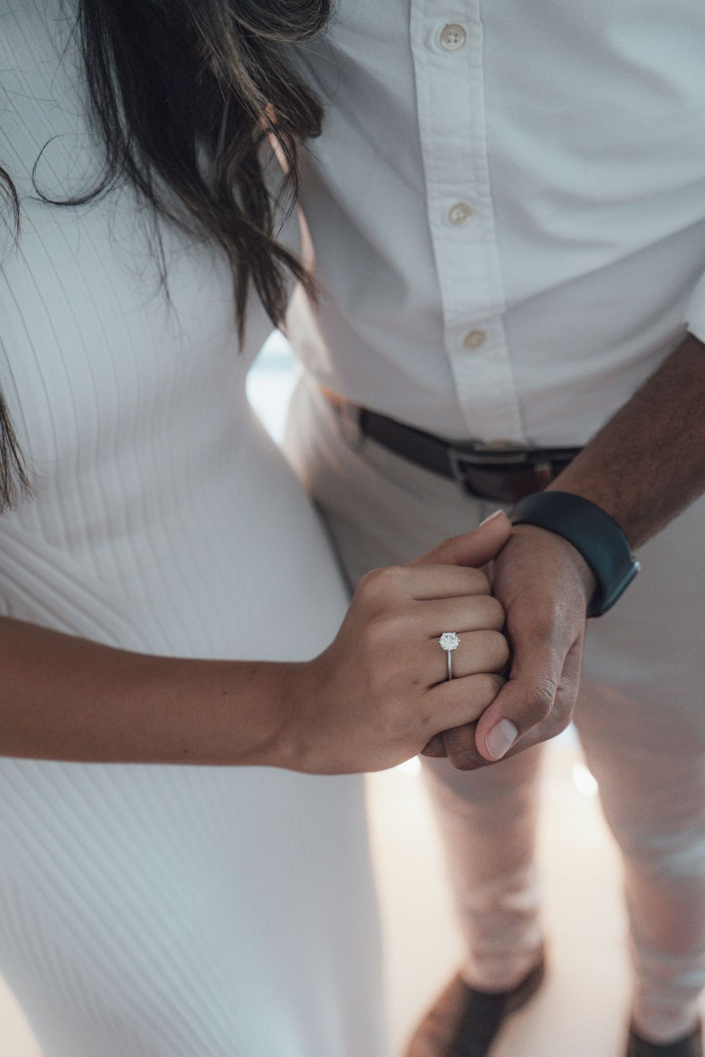 Close-up of hands wearing engagement ring with white button-down shirt in soft lighting.