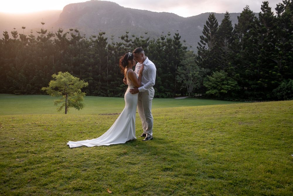Newlyweds embrace on grassy field with mountain landscape at sunset.