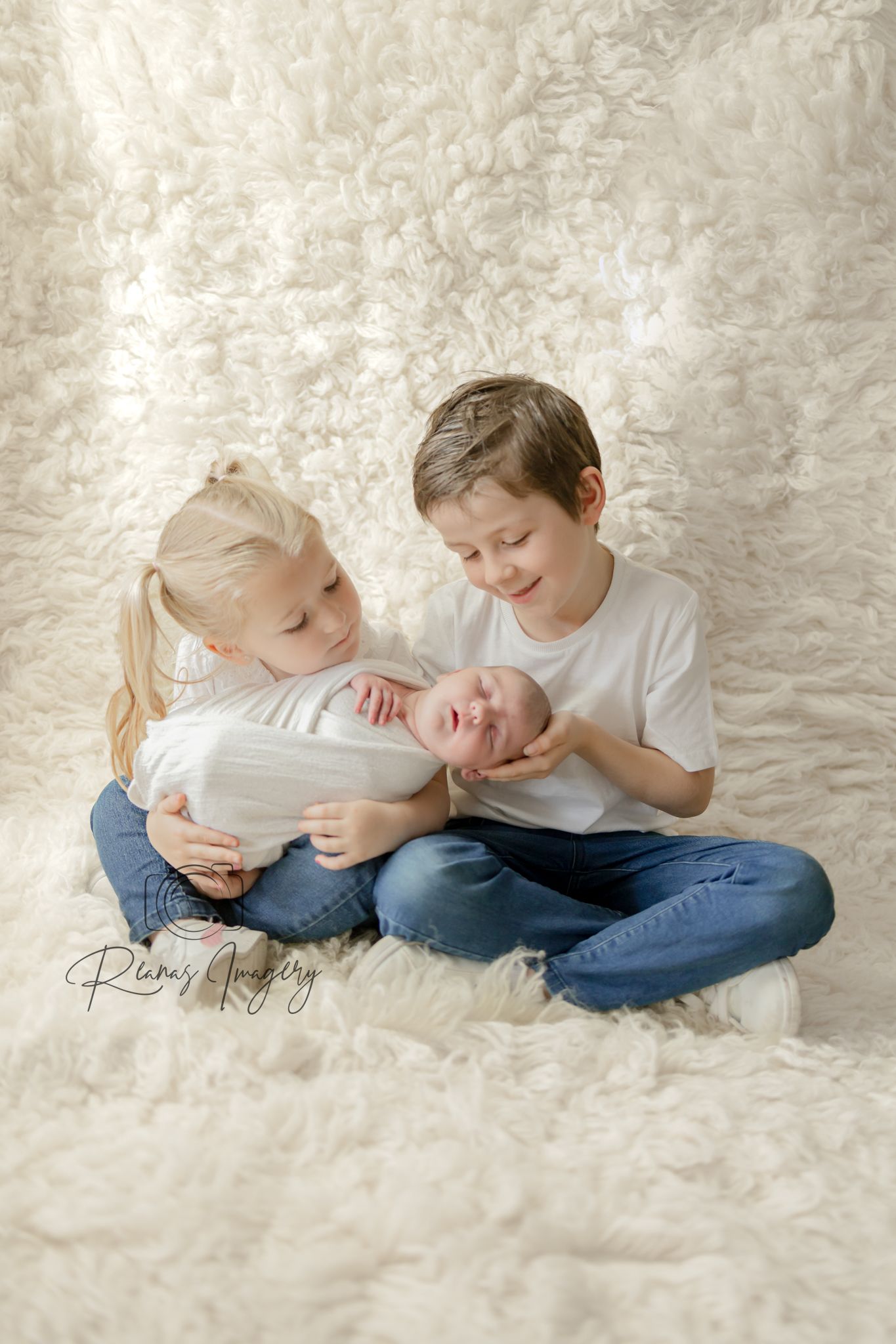 Two older siblings in white shirts and jeans tenderly holding a sleeping newborn on a fluffy white backdrop.