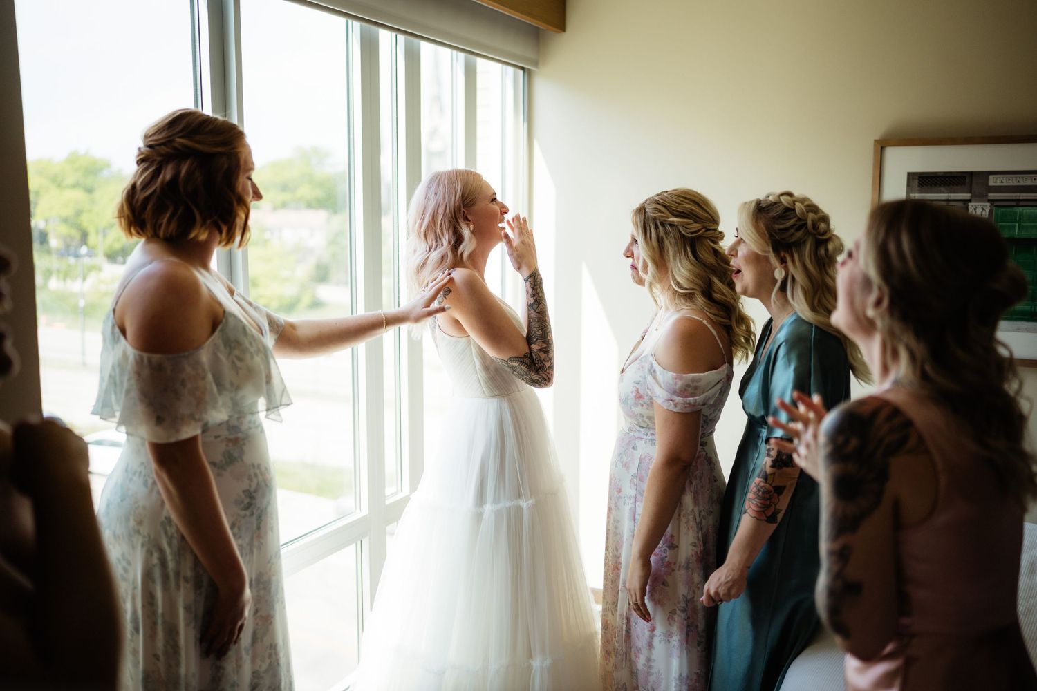 A group in white dresses gather by large windows in a sunlit room.