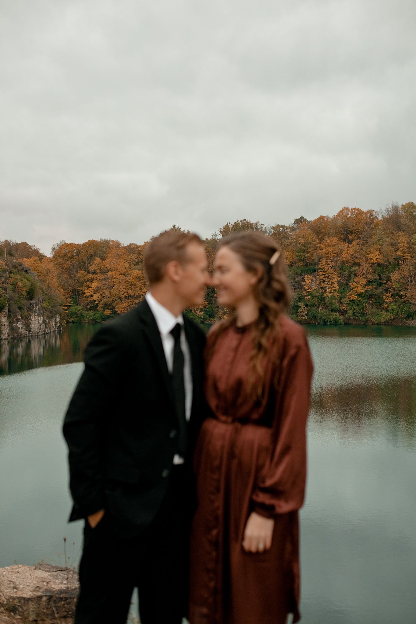 Romantic autumn engagement photos by a lake with fall foliage reflected in the calm waters.