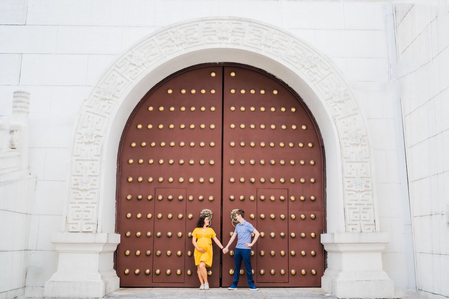Two people in yellow and blue outfits stand against a large ornate brown door.