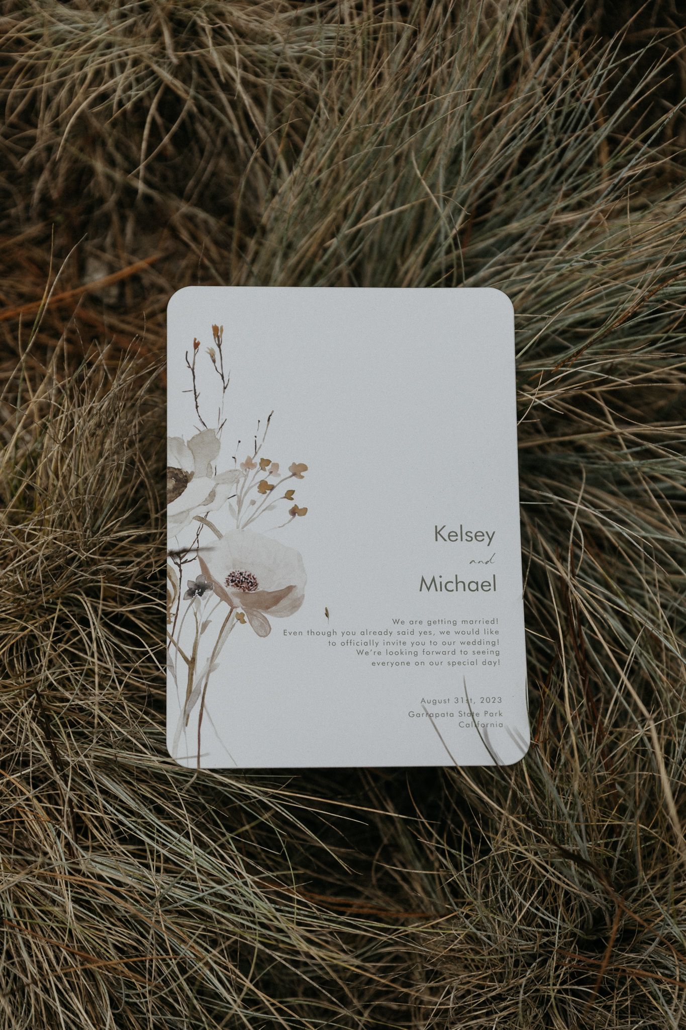 A minimalist wedding invitation with delicate floral illustrations rests on dried grass.