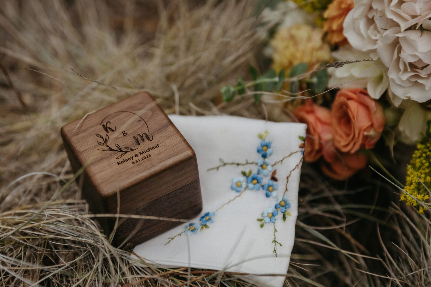 Wooden ring box and wedding stationery with blue floral details nestled in dried grass.