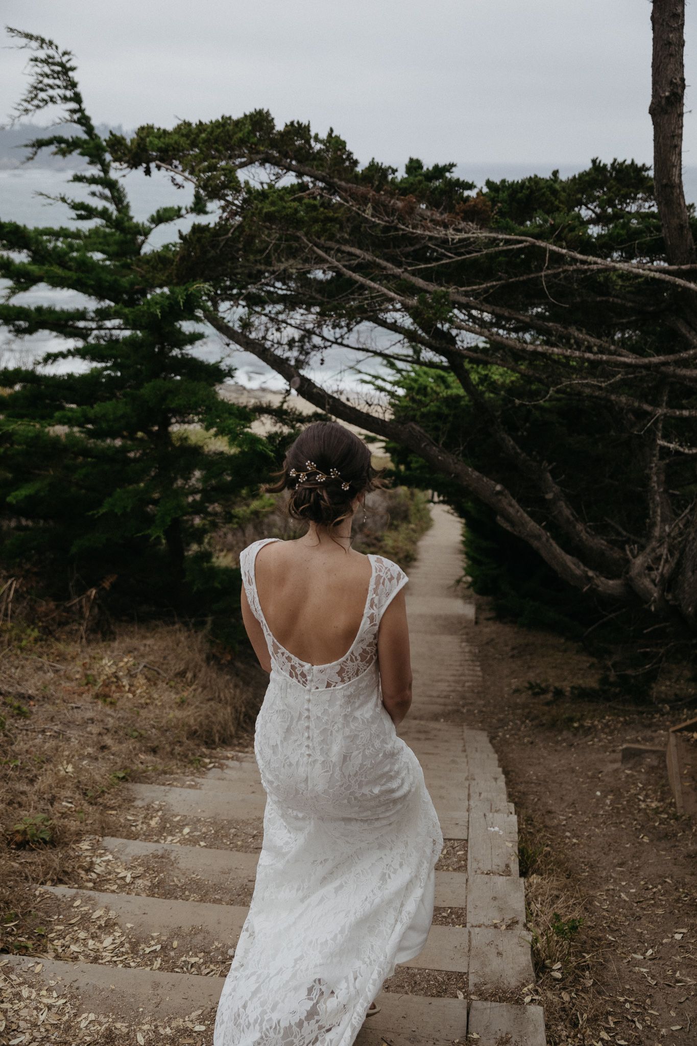 Lace wedding dress photographed from behind on stone stairs surrounded by coastal trees.