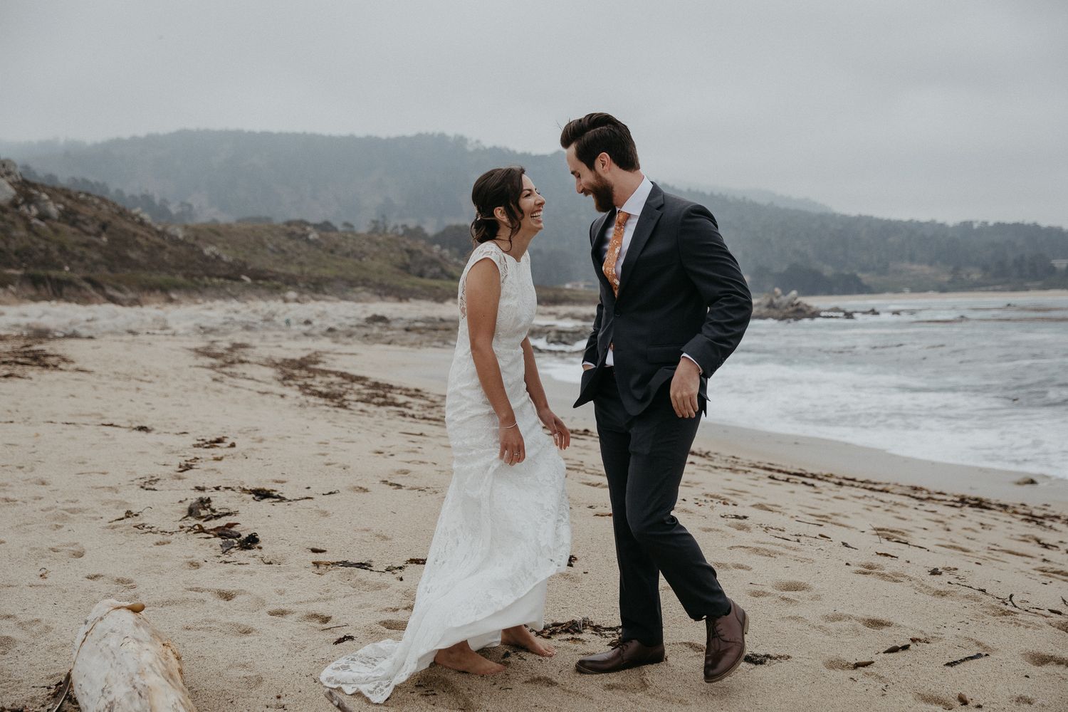 Couple in wedding attire walking together on a misty beach.