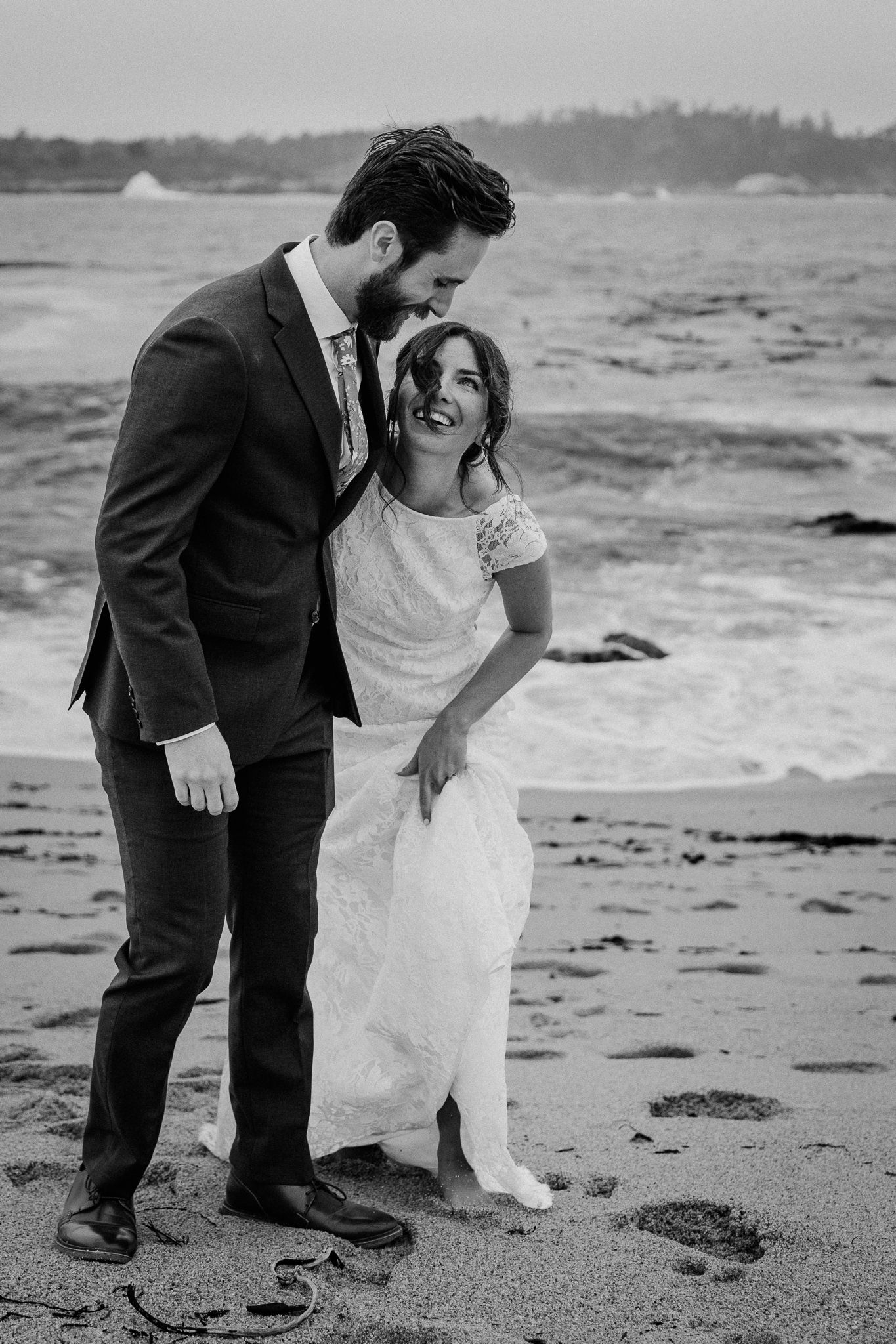 A romantic beach wedding photo captures a couple embracing and laughing in formal attire on a sandy shoreline.