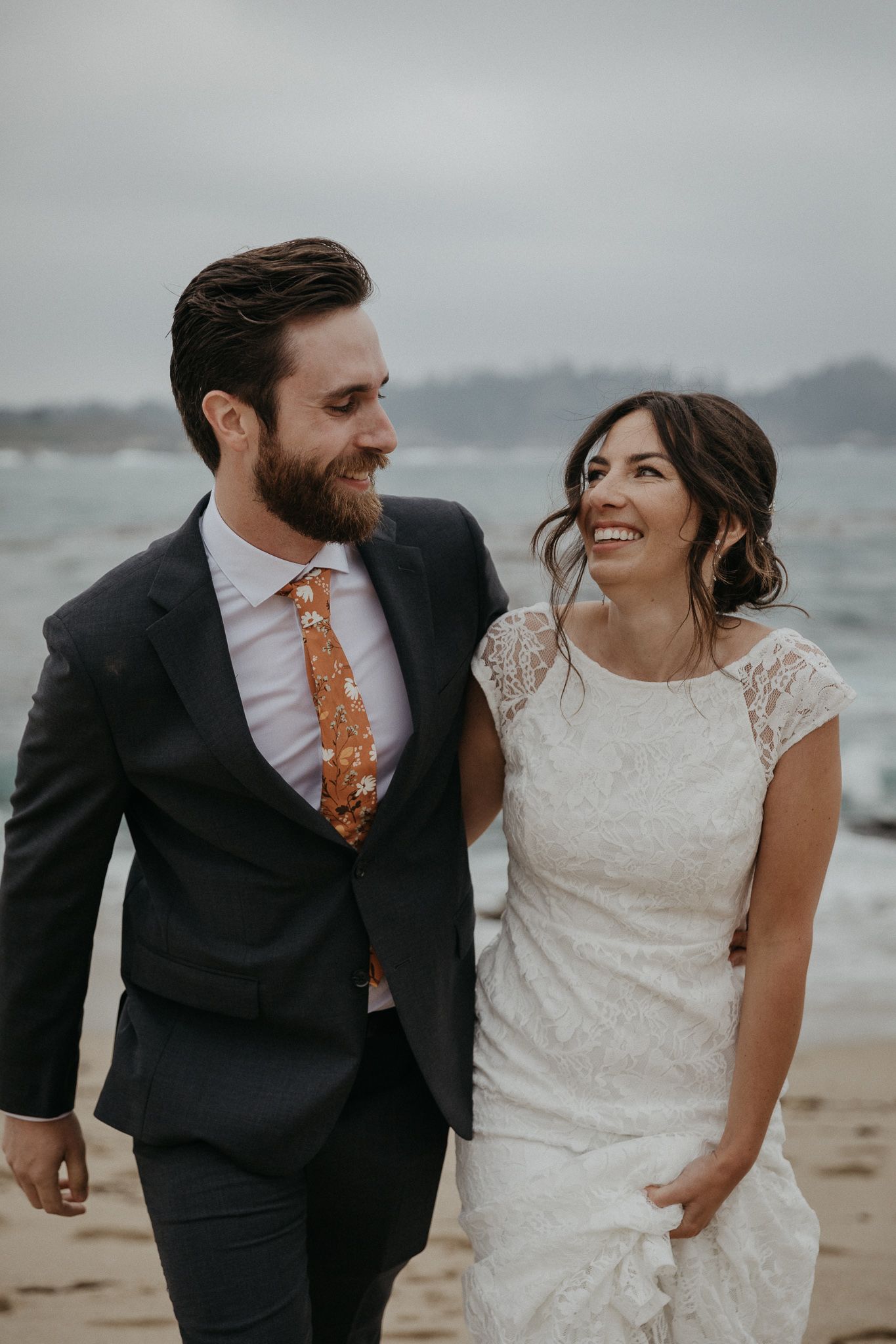 A beachside wedding portrait shows a couple in formal attire smiling and walking together on the sand.