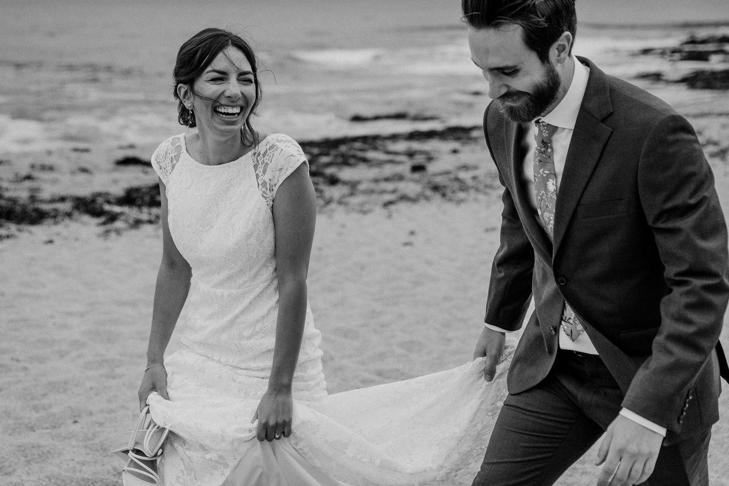 A candid black and white moment of a couple adjusting the wedding dress on a beach.