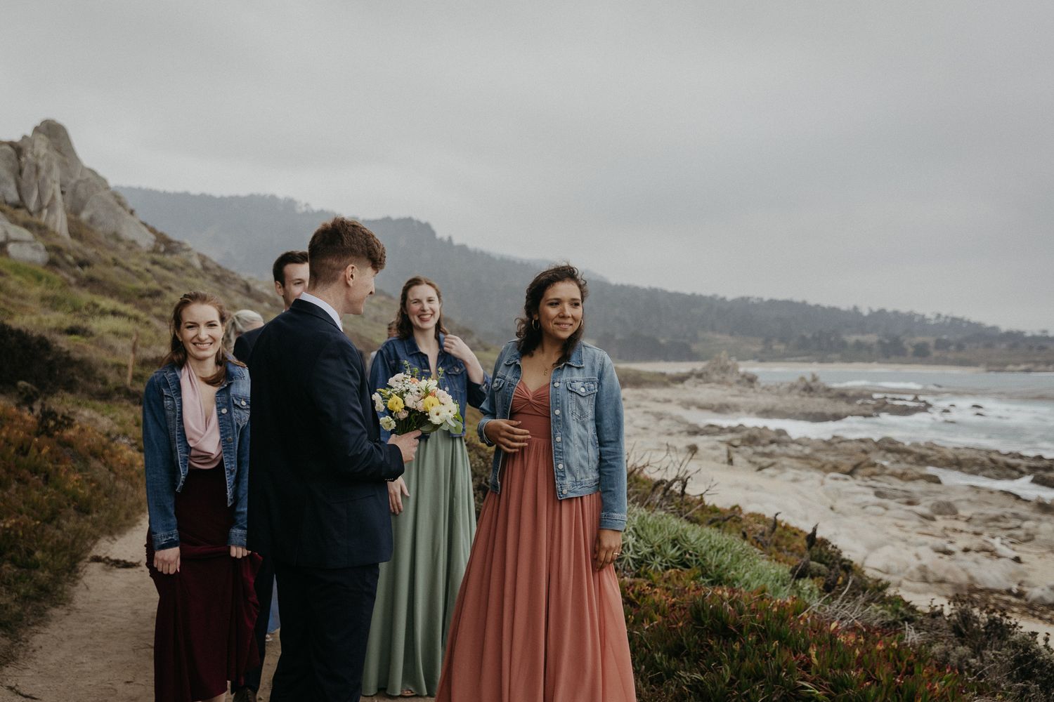 Wedding party members in colorful dresses pose along a scenic coastal cliff path.