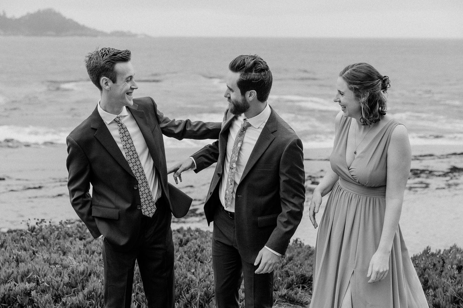 A black and white photo of wedding party members sharing a lighthearted moment by the ocean.