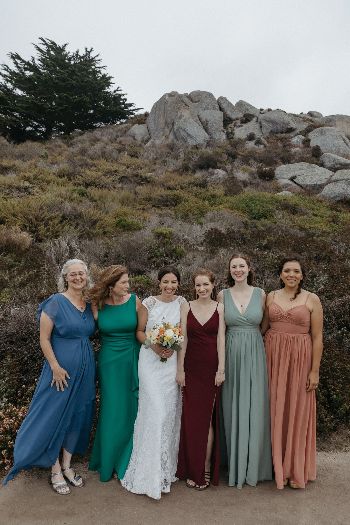 Wedding party members in coordinated dresses pose against a rocky coastal backdrop.