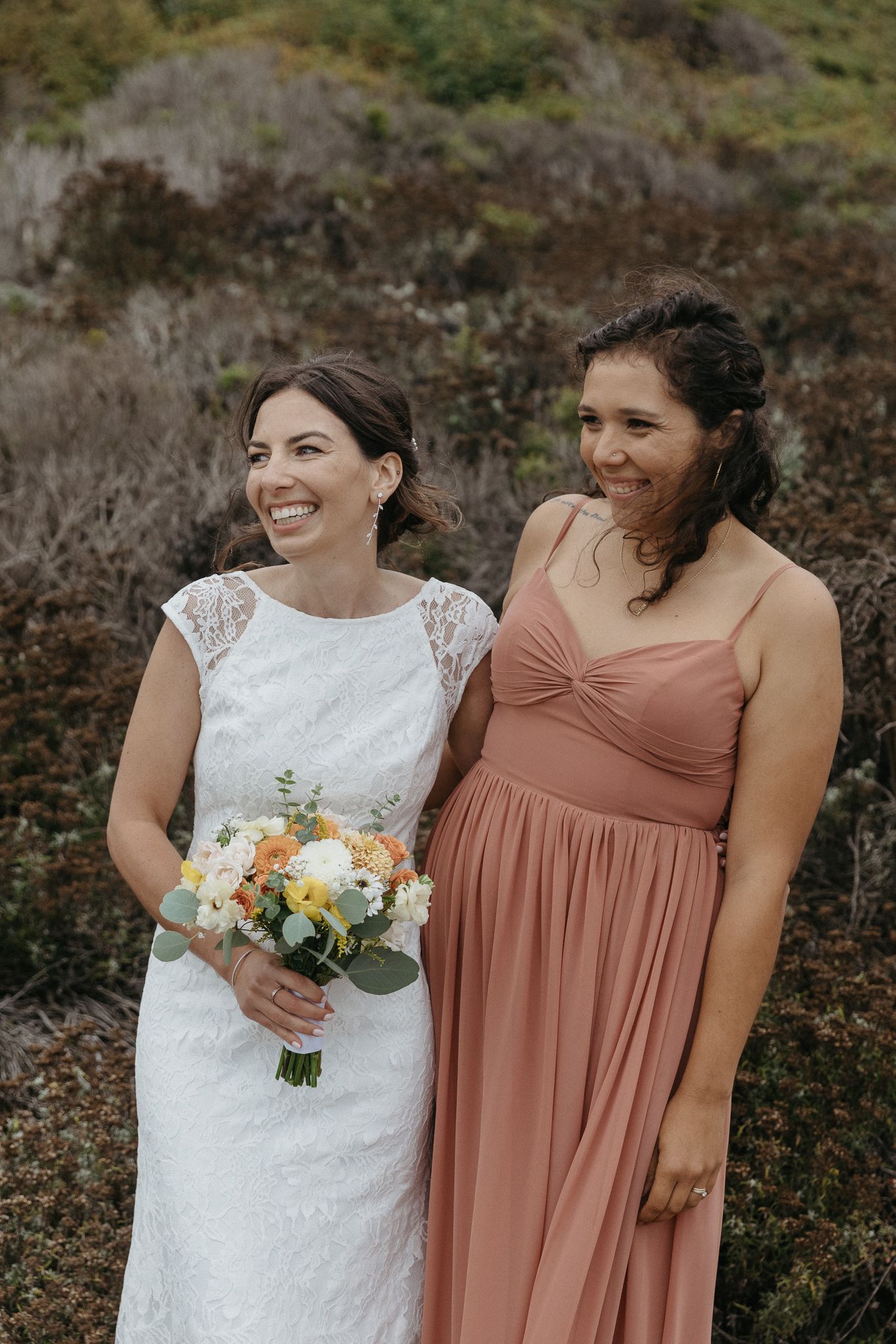 A bride with floral bouquet stands with bridesmaid in peach dress against natural coastal backdrop.