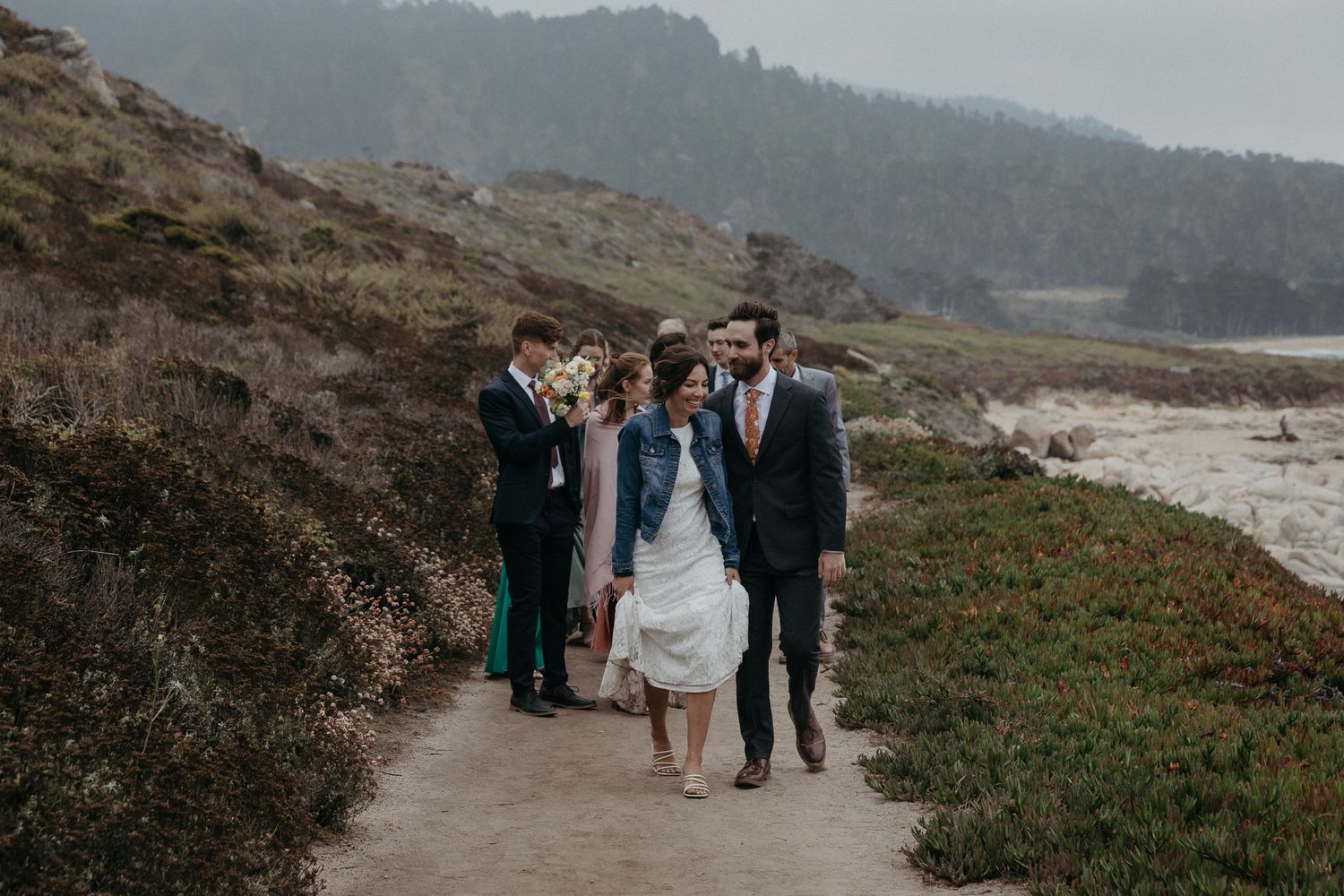Wedding party walks along a coastal path with mountains and ocean views in the background.