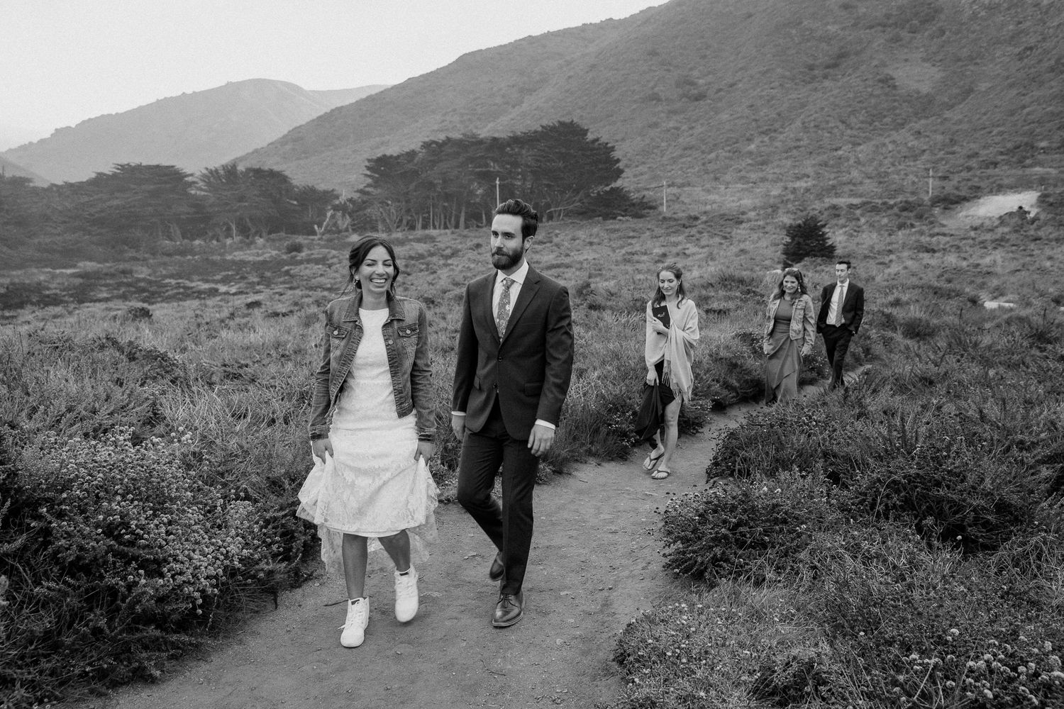 A couple walks along a coastal trail with mountains in the background.