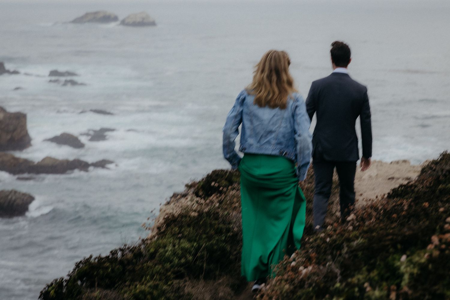 A couple stands on a cliff overlooking a misty ocean with rocks visible below.