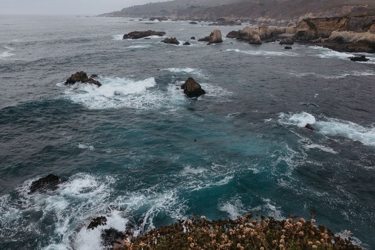 Dramatic coastline view of Big Sur with turquoise waters crashing against rocky outcrops on a cloudy day.