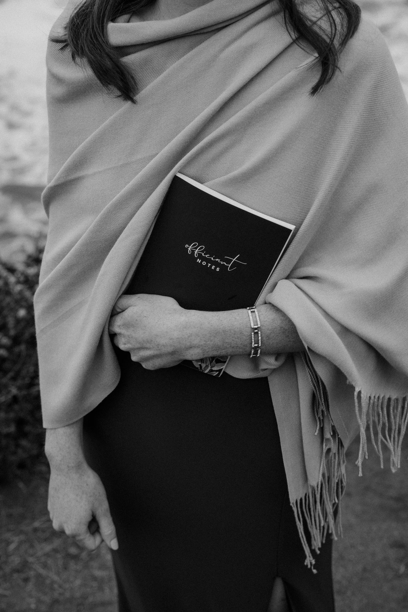 Someone holds a black ceremony book while wearing a gray shawl with fringe detail.