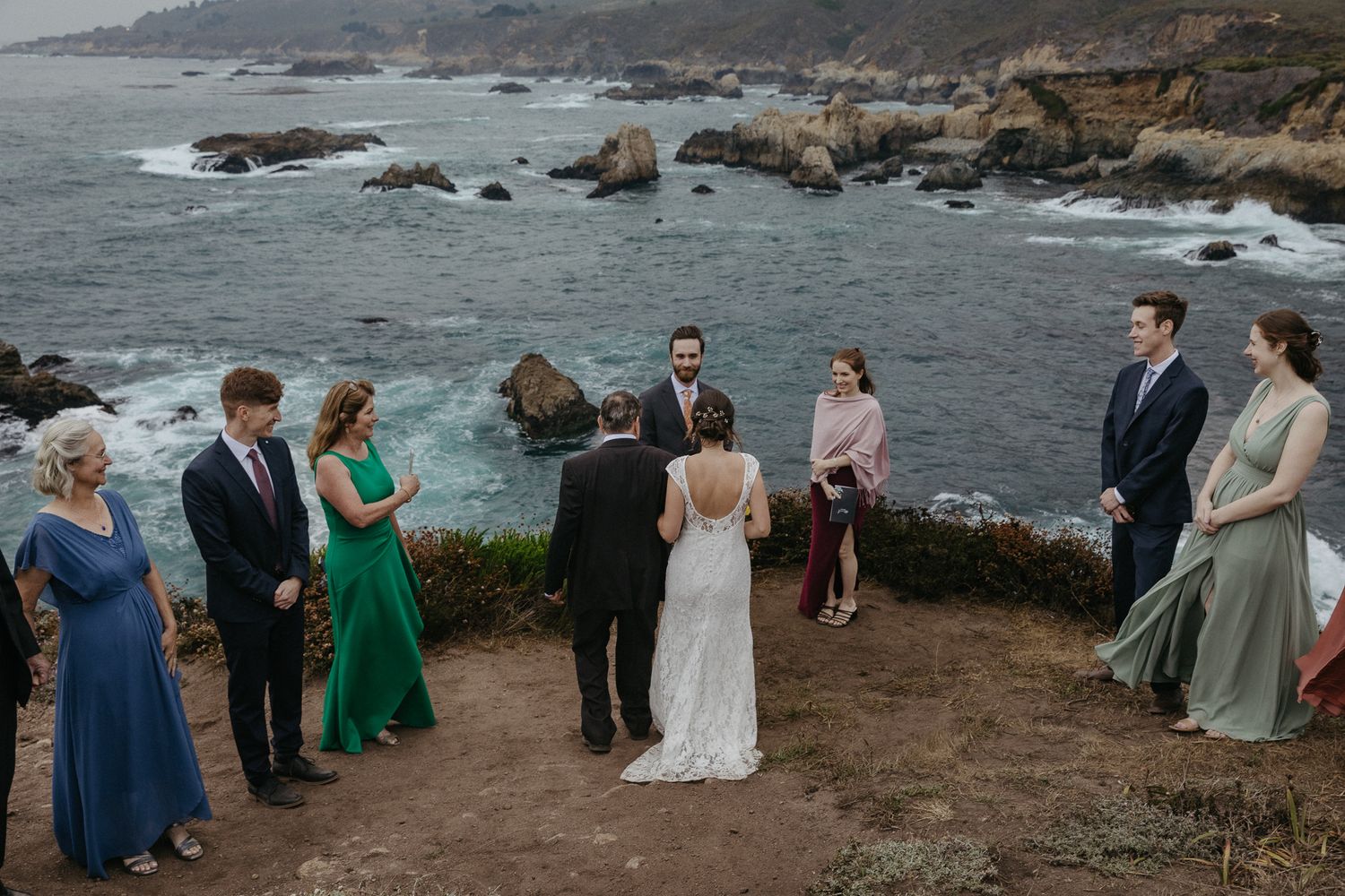 Small wedding gathering on coastal cliff with ocean and rocky shoreline in background.