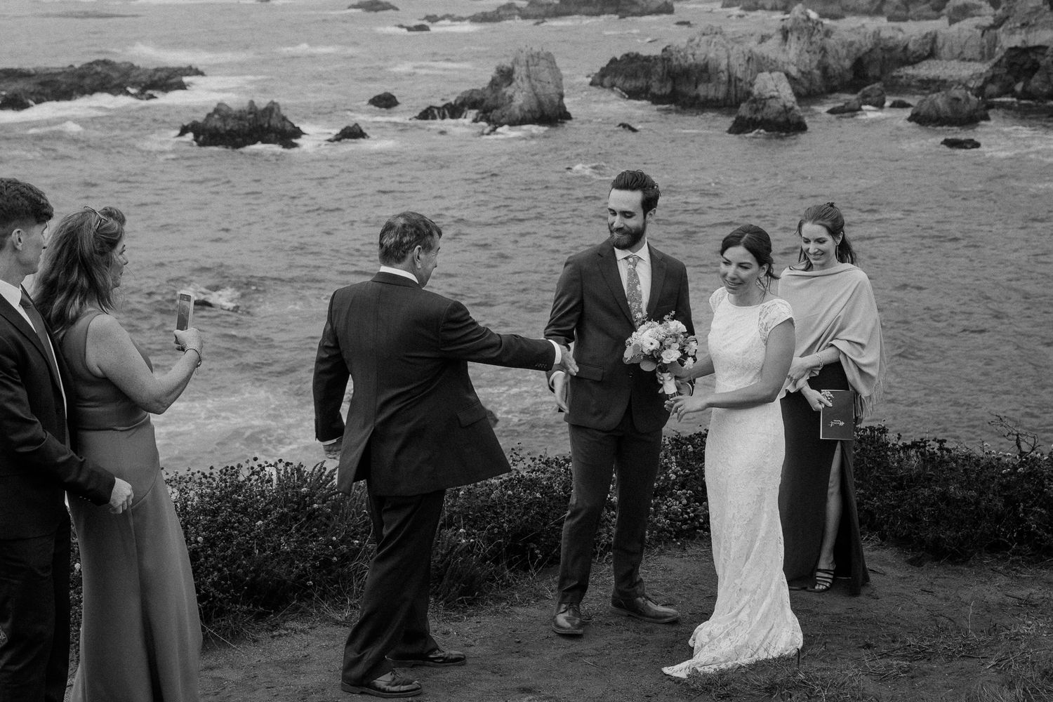 Black and white photo of wedding celebration on scenic coastal overlook.