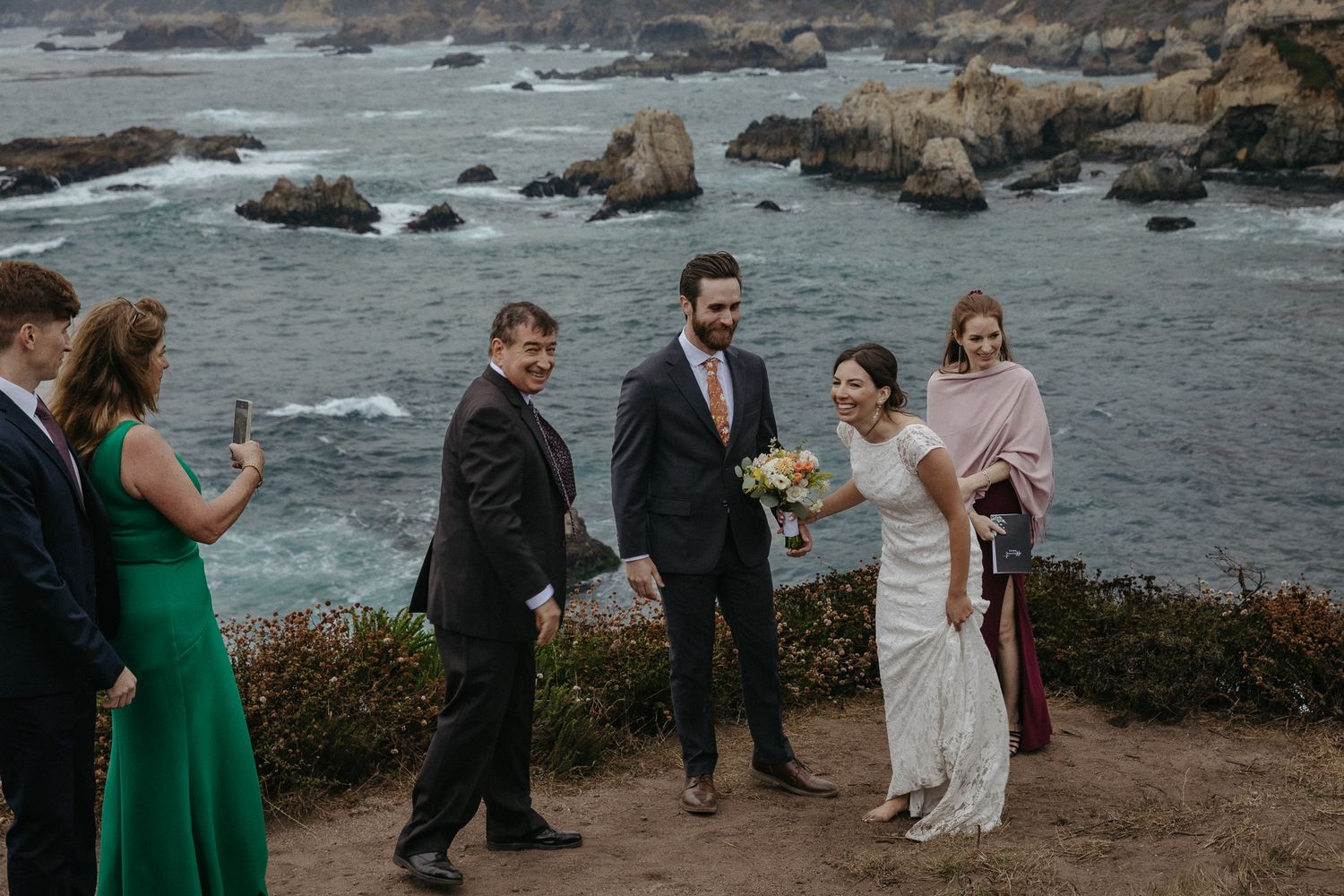 Wedding party celebrating on cliff edge with dramatic Pacific coast backdrop.