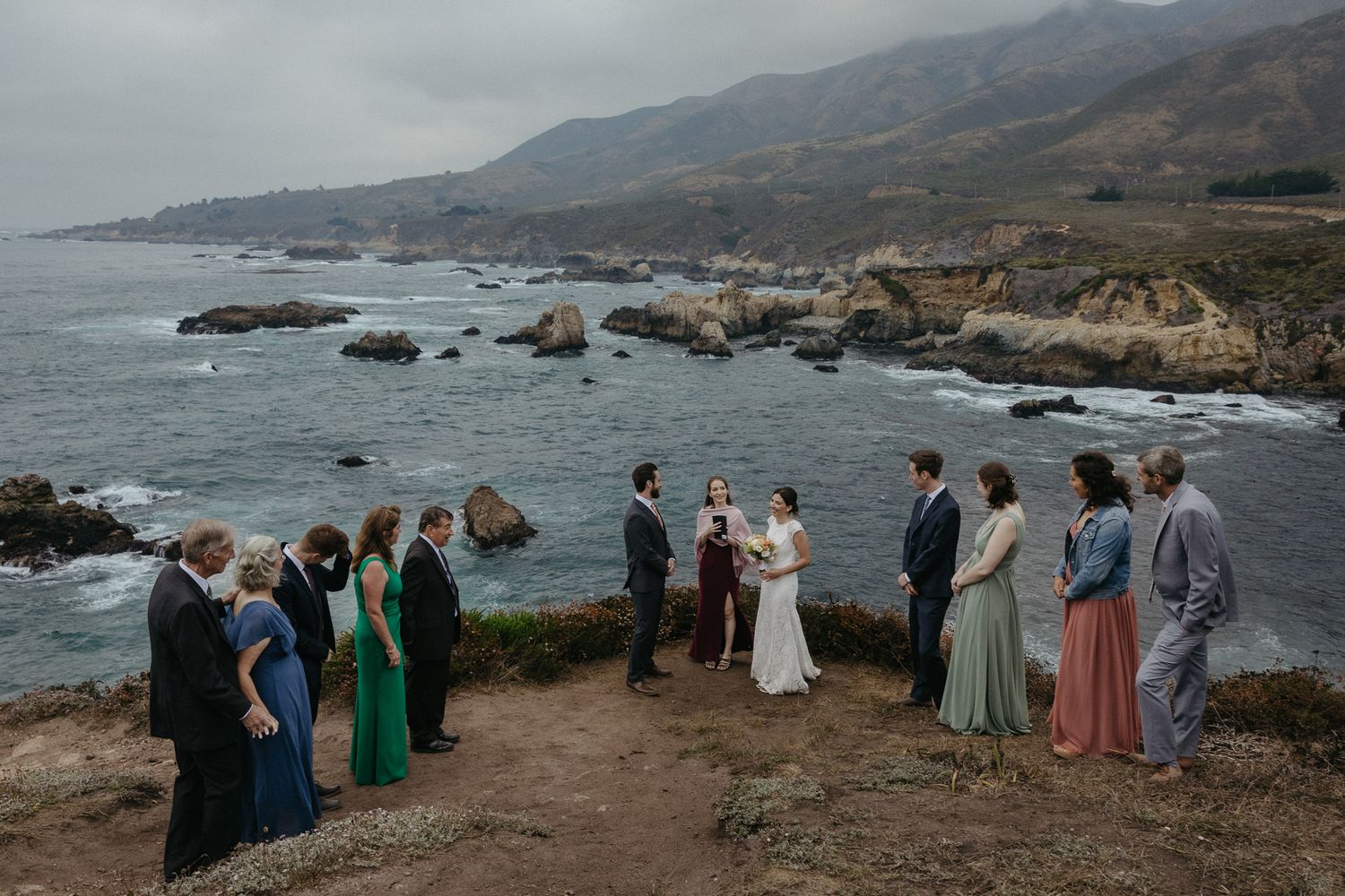 Intimate wedding ceremony group gathered on coastal bluff under cloudy skies.