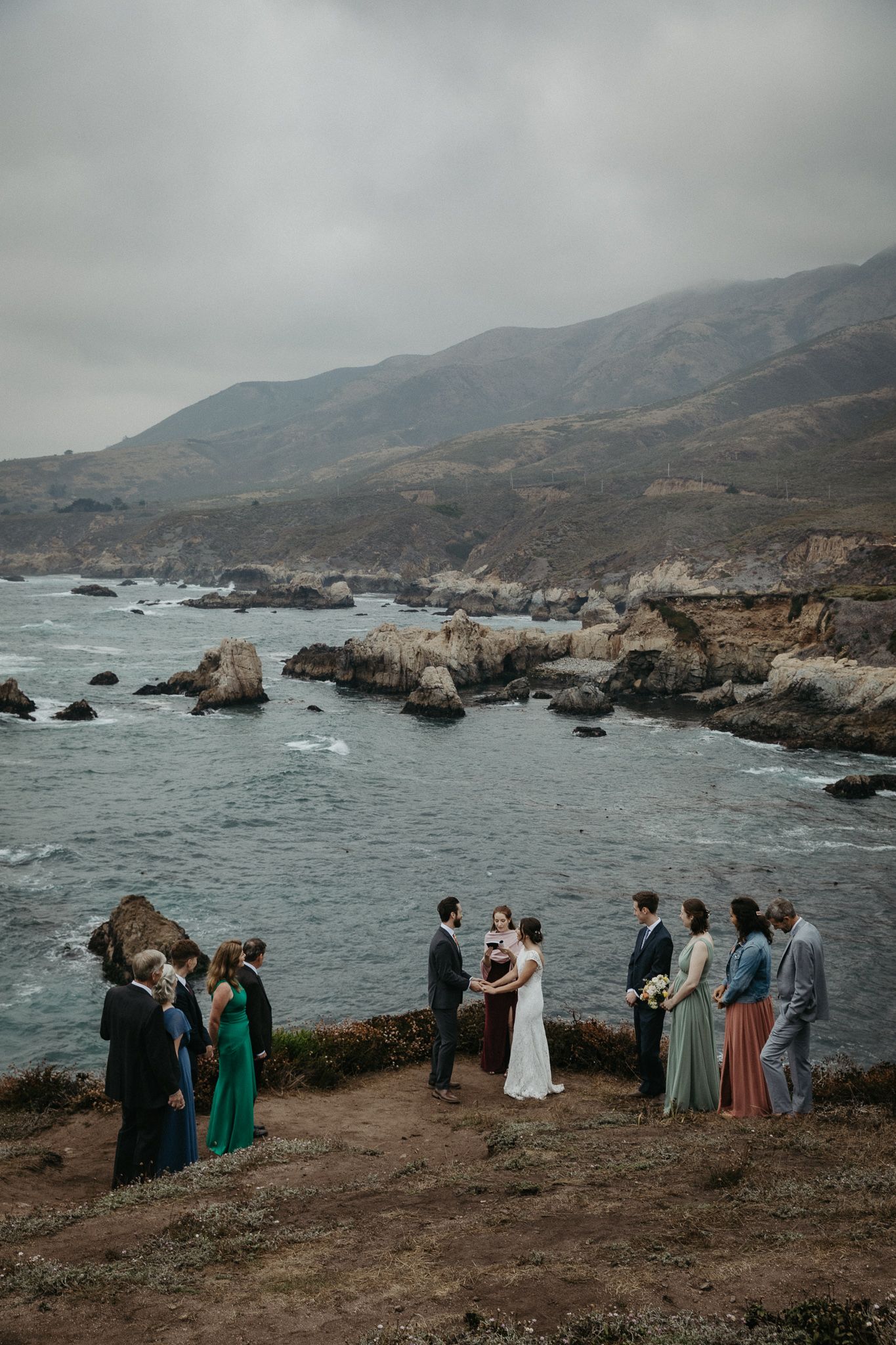 Intimate wedding ceremony on dramatic coastal cliff with mountain and ocean backdrop.