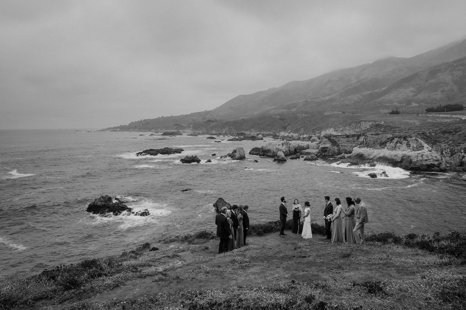 Black and white landscape of ceremony group on coastal cliffs with mountain backdrop.