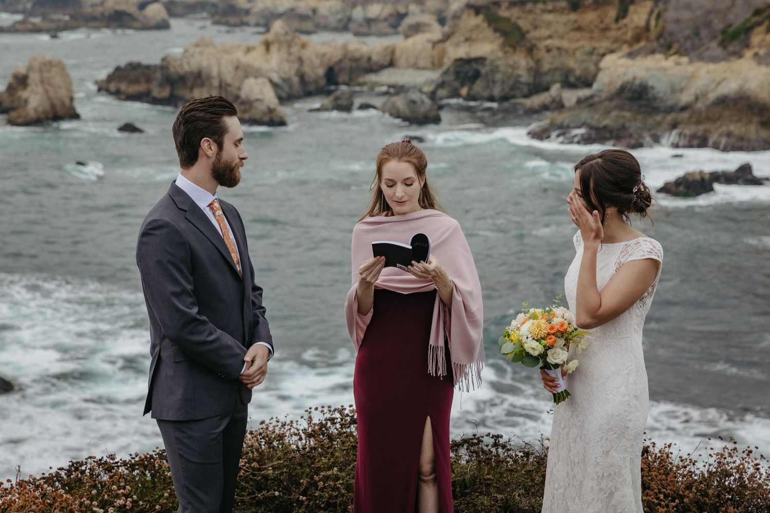 An intimate coastal wedding ceremony takes place on a cliff overlooking a rocky shoreline.