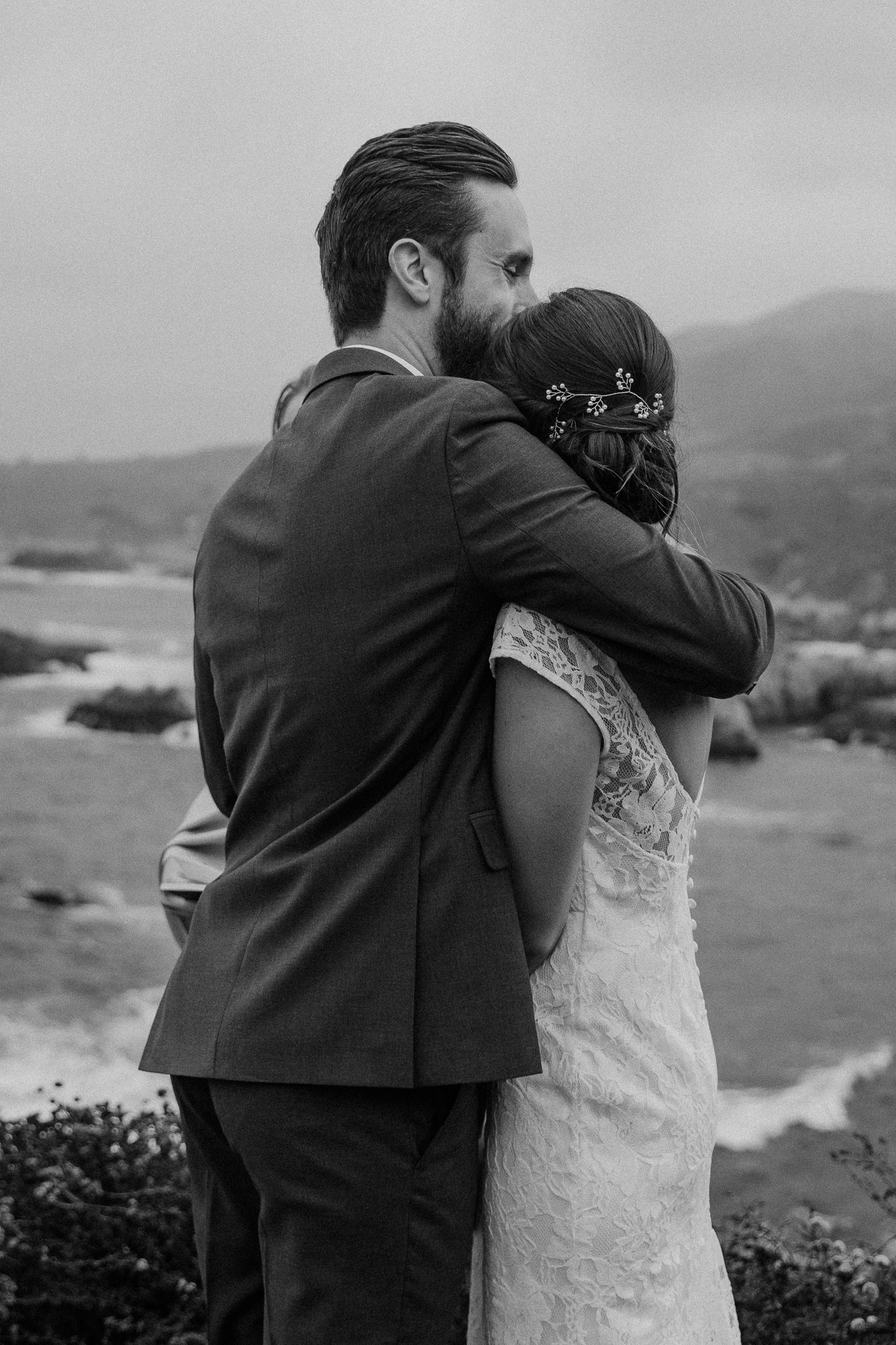 A romantic embrace captured in black and white against a dramatic coastal landscape.