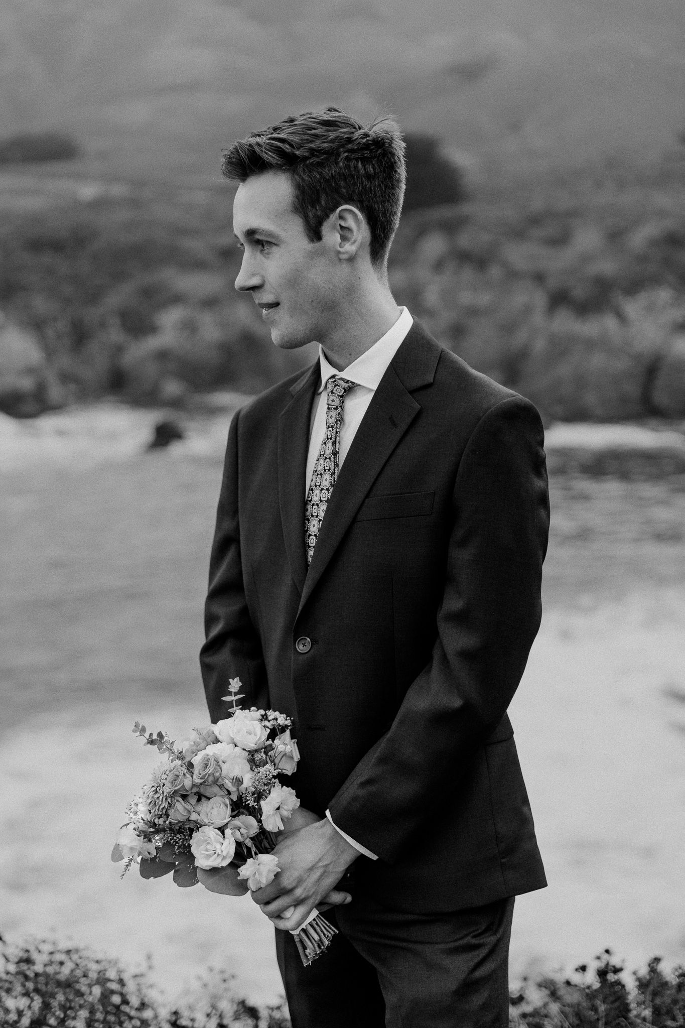 A groom in a dark suit holds a delicate bouquet against a mountainous backdrop.