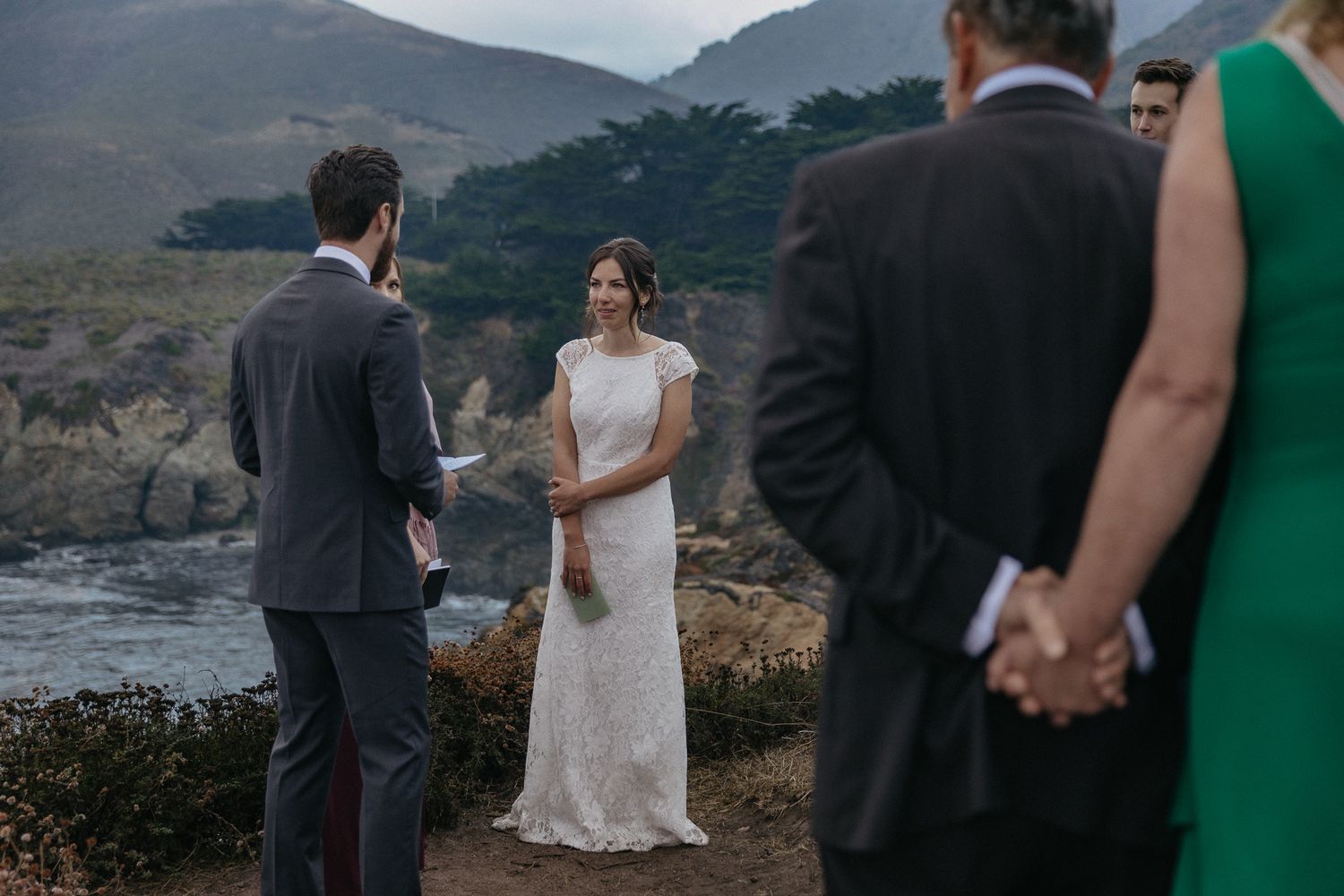 A wedding ceremony unfolds on a scenic coastal cliff with mountains in the background.