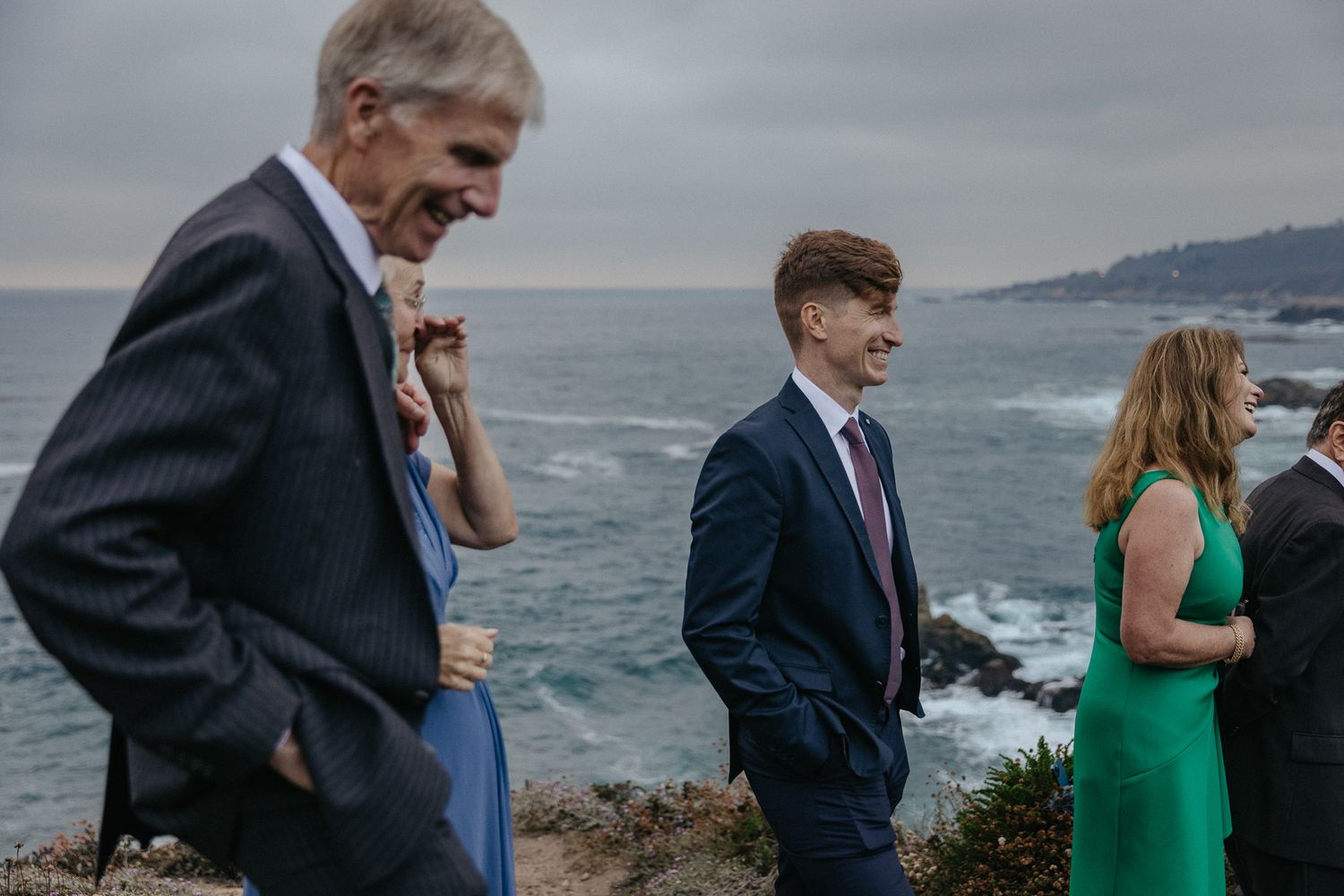 Wedding guests gather on a coastal overlook during an overcast ceremony.