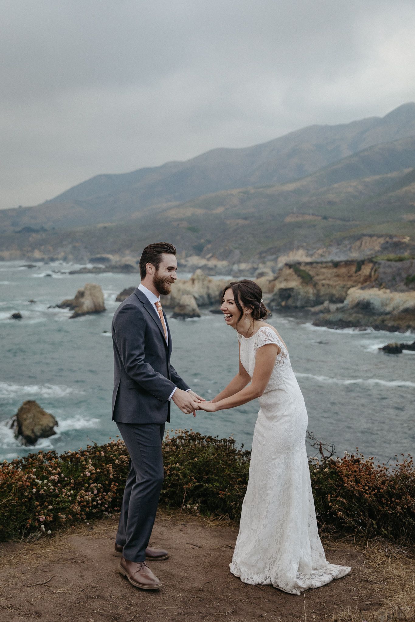 Wedding couple standing on coastal cliff with dramatic ocean and mountain backdrop.