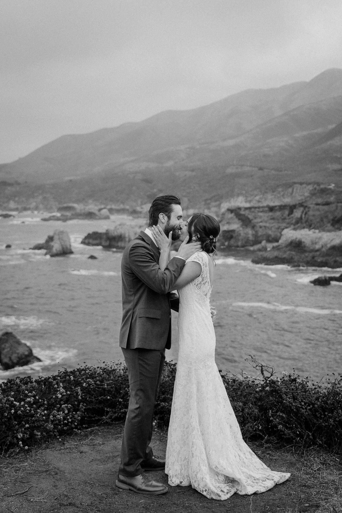 Romantic black and white portrait of wedding couple embracing on clifftop overlooking ocean.