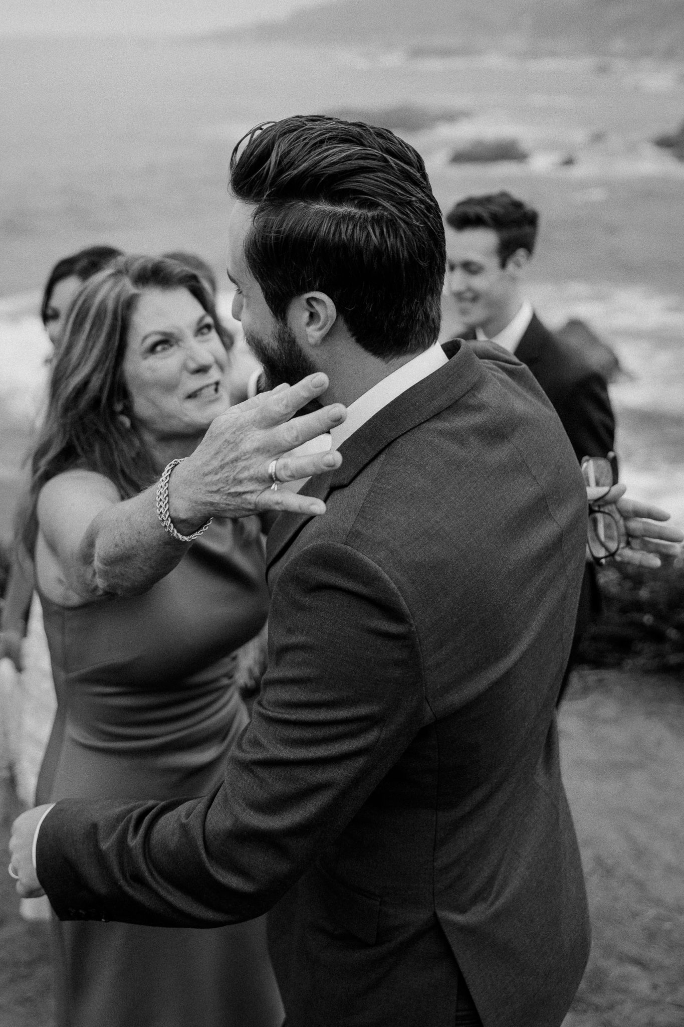 Candid moment of wedding guests sharing embraces against coastal backdrop.